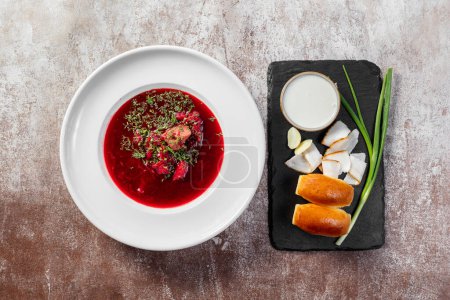 Photo for Ukrainian borscht soup with garlic donuts, green onions and bacon, next to it is a bowl of sour cream. The soup is poured into a light ceramic plate. Dishes stand on a light background. - Royalty Free Image