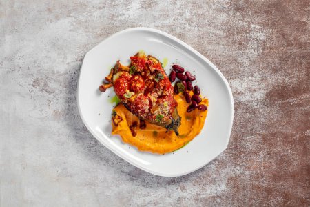 Photo for Eggplant baked with tomatoes and cheese in a spicy paprika sauce with chanterelle mushrooms and nuts. Next to pumpkin puree and red beans. Food on a light ceramic plate. The plate stands on a light background. - Royalty Free Image