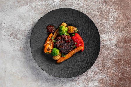 Photo for Grilled filet mignon steak with vegetables carrots, paprika broccoli in cherry sauce. Food lies on a dark slate plate on a light background. - Royalty Free Image
