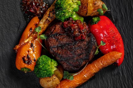 Foto de Filete a la parrilla mignon steak con verduras zanahorias, brócoli pimentón en salsa de cereza. La comida se encuentra en una placa de pizarra oscura sobre un fondo claro. - Imagen libre de derechos