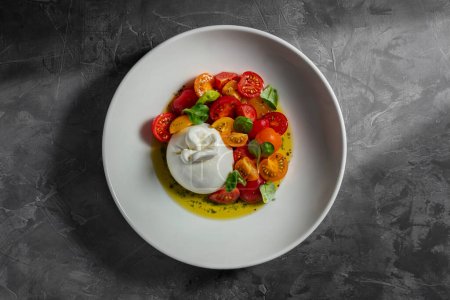 Téléchargez les photos : Mozzarella aux tomates cerises jaunes et rouges et feuilles de basilic dans de l'huile d'olive dans une assiette en céramique blanche. La plaque se tient sur un fond gris. - en image libre de droit