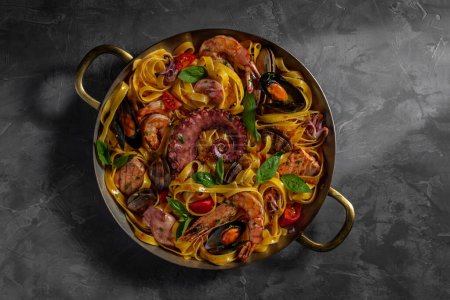 Photo for Homemade pasta with shrimp, octopus, mussels, squid, cherry tomatoes and basil leaves in a round handle pan. The frying pan stands on a gray background. - Royalty Free Image