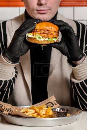 Téléchargez les photos : Burger avec galette de boeuf, sauce au fromage avec paprika et laitue. A proximité se trouvent des frites et un bateau à sauce au ketchup. Un homme tient un hamburger dans ses mains et prend une bouchée. - en image libre de droit