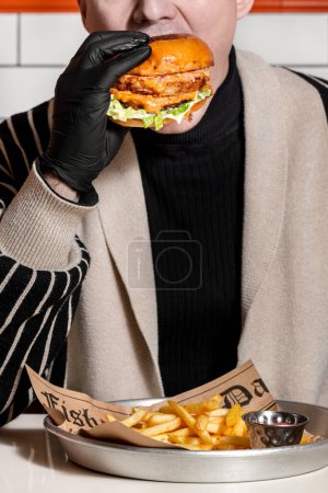 Téléchargez les photos : Burger avec galette de boeuf, sauce au fromage avec paprika et laitue. A proximité se trouvent des frites et un bateau à sauce au ketchup. Un homme tient un hamburger dans ses mains et prend une bouchée. - en image libre de droit