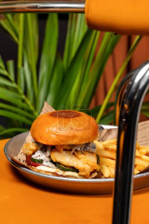 Téléchargez les photos : Burger avec filet de perche, cornichons, sauce aux champignons crémeuse et morceaux d'anguille fumée. À proximité se trouvent des frites et un bol de sauce au ketchup. La nourriture est dans un bol en métal. - en image libre de droit