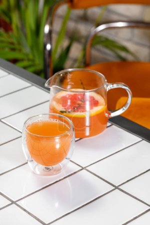 Téléchargez les photos : Le thé aux agrumes avec du thym, des oranges rouges et du citron dans une théière transparente est versé dans une tasse transparente. Les plats se tiennent sur un fond clair. - en image libre de droit