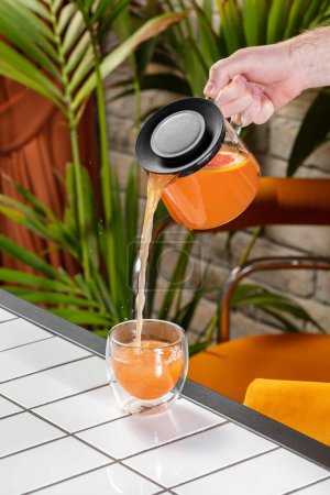 Téléchargez les photos : Le thé aux agrumes avec du thym, des oranges rouges et du citron dans une théière transparente est versé dans une tasse transparente. Les plats se tiennent sur un fond clair. - en image libre de droit