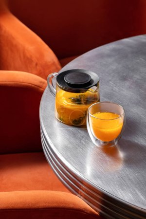 Téléchargez les photos : Thé d'argousier au thym et citron dans une théière transparente avec couvercle. À proximité se trouve une tasse de thé transparente. Les plats sont sur une table en métal, chaises orange sont à proximité. - en image libre de droit