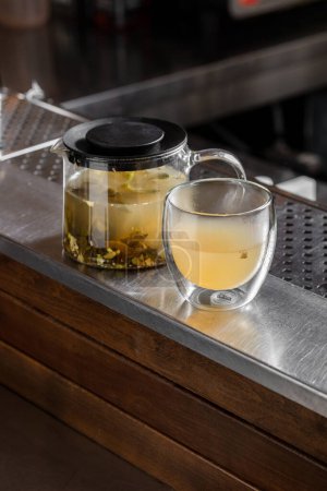 Foto de Té de menta con limón, canela y hojas de menta en una tetera transparente con tapa. Cerca hay un vaso de té. Los platos están en un soporte de metal. - Imagen libre de derechos