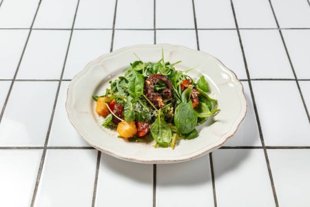 Téléchargez les photos : Salade chaude avec perche panée, poulpe, tomates, parmesan et laitue. La nourriture se trouve dans une plaque de céramique légère. - en image libre de droit