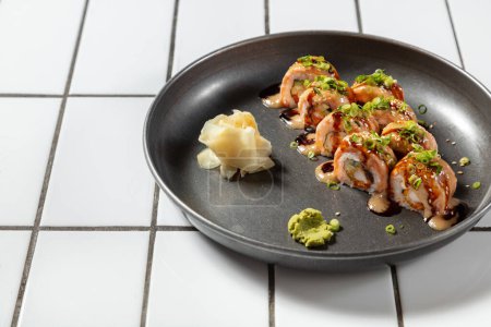 Foto de Sushi con salmón, tempura de camarones y aguacate. Cubierto con salsa unagi y mayonesa de chile japonés. Adornada con pequeños aros de cebolla verde. Sushi se encuentra en una placa de metal sobre un fondo claro. - Imagen libre de derechos