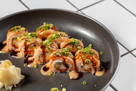 Foto de Sushi con salmón, tempura de camarones y aguacate. Cubierto con salsa unagi y mayonesa de chile japonés. Adornada con pequeños aros de cebolla verde. Sushi se encuentra en una placa de metal sobre un fondo claro. - Imagen libre de derechos