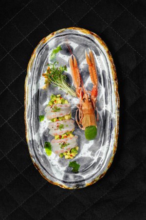 Foto de Langoustine sashimi encima del tartar de aguacate con rodajas de chile y cebolletas encima. Cerca se encuentra la cáscara y microverde. La comida se encuentra en un plato de cerámica con un patrón sobre un fondo negro. - Imagen libre de derechos