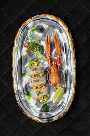 Photo for Langoustine sashimi on top of avocado tartare with chili slices and scallions on top. Nearby lies shell and microgreen. Food lies on a ceramic plate with a pattern on a black background. - Royalty Free Image