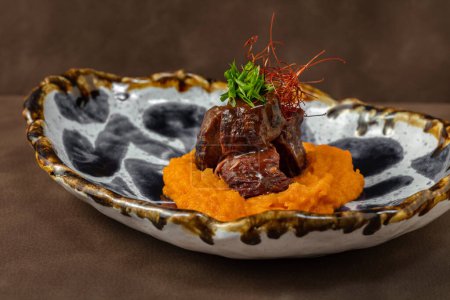Photo for Veal cheeks stewed in red wine on top of pumpkin puree. Chopped green onions are on top of the cheeks. The food lies in a ceramic plate with kzor. The plate stands on a brown, leather background. - Royalty Free Image