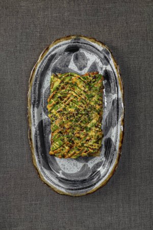 Photo for Corn tortilla toast with eel in unagi sauce and avocado slices. Top with chilli-paprika sauce. Food lies on a ceramic plate with a pattern on a gray fabric background. - Royalty Free Image