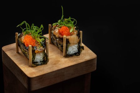 Foto de Vieira gunkan con arroz y caviar tobiko en algas nori. Gunkans de pie sobre un soporte de madera sobre un fondo negro. - Imagen libre de derechos