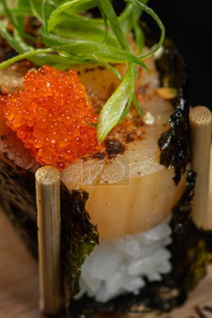 Téléchargez les photos : Gunkan de pétoncle avec riz et caviar tobiko dans les algues nori. Gunkans debout sur un support en bois sur un fond noir. - en image libre de droit