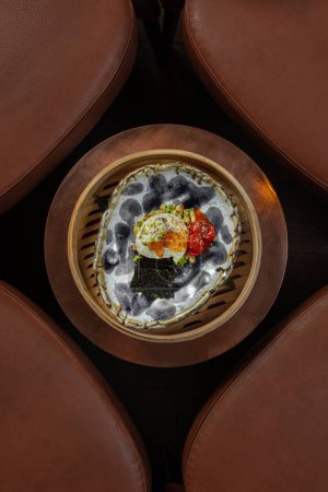 Photo for Charcoal-smoked mozzarella with avocado tartare, pickled paprika and nori seaweed in a patterned ceramic plate. The plate is in a wooden dish on a metal table. - Royalty Free Image