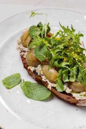 Photo for Forshmak of herring on buckwheat toast with pickled grapes, mint leaves and cilantro. Food lies on a light ceramic plate. The plate stands on a light background. - Royalty Free Image