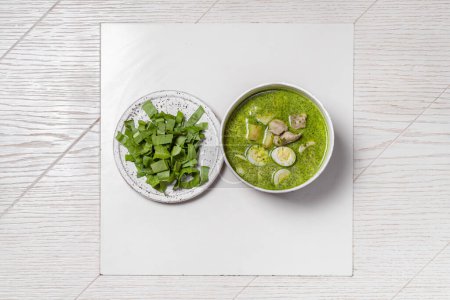 Photo for Green borscht with pieces of veal, potatoes, spinach and quail eggs. The soup is poured into a light ceramic plate, next to it lies a lid with greenery, there is a bottle and a glass of white wine. - Royalty Free Image