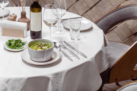 Photo for Green borscht with pieces of veal, potatoes, spinach and quail eggs. The soup is poured into a light ceramic plate, next to it lies a lid with greenery, there is a bottle and a glass of white wine. - Royalty Free Image