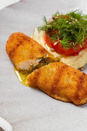 Photo for Chicken Kiev cutlet breaded with butter and herbs inside, mashed potatoes, tomato and microgreens on top. Food lies in a light ceramic plate on a light background. - Royalty Free Image