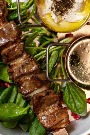 Photo for Lamb shish kebab on a metal skewer with pieces of onion. The meat lies on top of the lettuce leaves. Nearby are two saucepans with different sauces in a metal plate. - Royalty Free Image