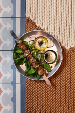 Téléchargez les photos : Agneau shish kebab sur une brochette en métal avec des morceaux d'oignon. La viande se trouve sur les feuilles de laitue. A proximité se trouvent deux casseroles avec différentes sauces dans une assiette en métal. - en image libre de droit