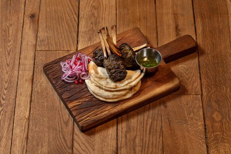 Photo for Rack of lamb wrapped in grape leaves and baked in the oven stands on portions of lavash. Nearby is a gravy bowl with oil and herbs and chopped red onion. Food on a wooden stand. - Royalty Free Image