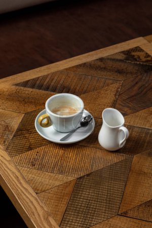 Téléchargez les photos : Café avec du lait dans une tasse en céramique légère sur une soucoupe avec une cuillère à café. A proximité se trouve une cruche à lait en céramique légère. Les plats sont sur une table en bois. - en image libre de droit