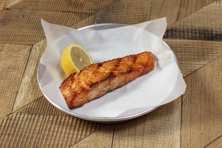Foto de Filete de salmón a la parrilla en salsa unagi. El pescado se encuentra sobre papel ligero en un recipiente redondo de metal. Cerca hay medio limón.. - Imagen libre de derechos