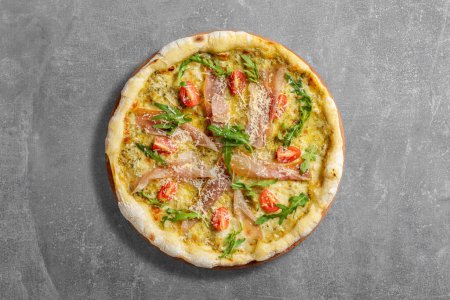 Téléchargez les photos : Pizza au levain avec morceaux de prosciutto, roquette, tomates cerises et parmesan râpé. Pizza repose sur un fond de pierre grise. - en image libre de droit