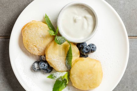 Photo for Cottage cheese pancakes with blueberries, mint leaves and powdered sugar on a light ceramic plate. Nearby is a gravy bowl with sour cream. The plate lies on a gray stone surface. - Royalty Free Image
