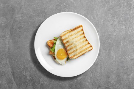 Foto de Sandwich con tostadas, salmón, huevo frito, hojas de albahaca y rúcula. El sándwich se encuentra en una placa de cerámica ligera sobre un fondo de piedra gris. - Imagen libre de derechos