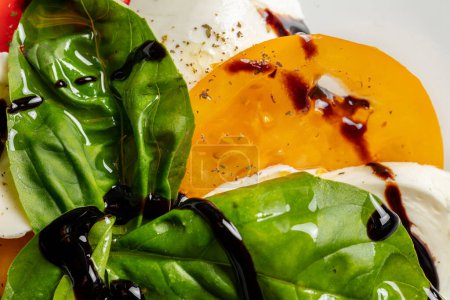Téléchargez les photos : Salade estivale de tomates rouges et jaunes avec mozzarella et feuilles de basilic avec vinaigrette balsamique. La salade repose sur une assiette en céramique légère sur un fond de pierre grise. - en image libre de droit