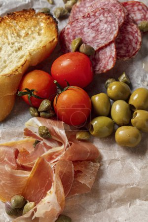 Téléchargez les photos : Amuse-gueule italien prosciutto, salami, olives, toasts à la baguette et tomates cerises reposent sur un parchemin léger sur une planche de bois. La planche repose sur un fond de pierre grise. - en image libre de droit