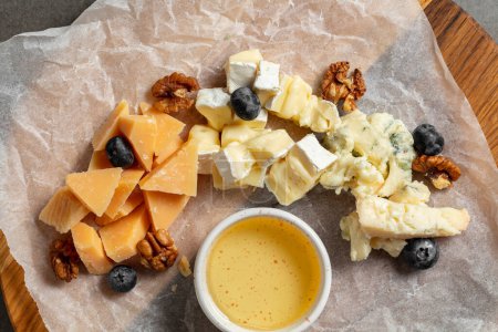Photo for Cheese plate of appetizers of brie, cheddar, dor blue and parmesan. Nuts and blueberries lie nearby and there is a bowl of honey. The food lies on a light parchment on a wooden board on a gray stone background. - Royalty Free Image