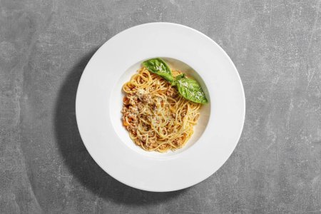 Foto de Espaguetis caseros con carne picada y salsa de tomate rematada con hojas de albahaca y queso parmesano rallado. Pasta se encuentra en una placa de cerámica ligera sobre un fondo de piedra gris. - Imagen libre de derechos