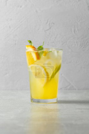 Téléchargez les photos : Un verre de limonade maison avec citron, citron vert et orange, glaçons et feuilles de menthe dans une tasse en verre transparent sur un fond de pierre grise. - en image libre de droit
