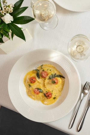 Photo for Ravioli with mozzarella and cherry tomatoes with grated parmesan on top. Food is in a light ceramic plate on a light tablecloth, there are several glasses of white wine and a vase of flowers nearby. - Royalty Free Image