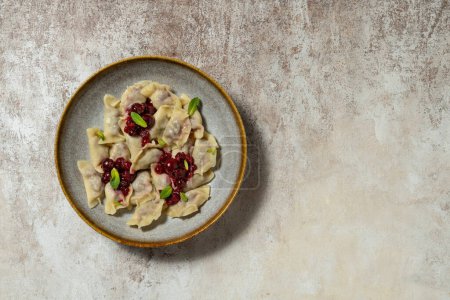 Photo for Ukrainian dish of dumplings with cherries and berries. Red currant and cherry sauce and mint leaves are on top. Food lies in a dark ceramic plate on a light fabric background. - Royalty Free Image