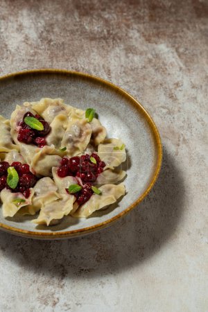 Photo for Ukrainian dish of dumplings with cherries and berries. Red currant and cherry sauce and mint leaves are on top. Food lies in a dark ceramic plate on a light fabric background. - Royalty Free Image