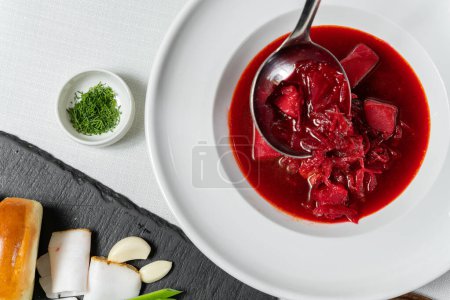 Téléchargez les photos : Soupe de bortsch ukrainienne dans une assiette en céramique ronde et légère. A proximité se trouve un stand d'ardoise avec des beignets à l'ail et des morceaux de saindoux. - en image libre de droit