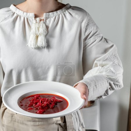 Téléchargez les photos : Soupe de bortsch ukrainienne dans une assiette en céramique ronde et légère. A proximité se trouve un stand d'ardoise avec des beignets à l'ail et des morceaux de saindoux. Une fille en ukrainien vyshyvanka tient une assiette dans ses mains. - en image libre de droit