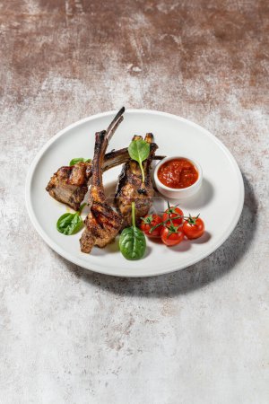 Photo for Three pieces of grilled lamb rack lie on a light ceramic plate. Nearby are cherry tomatoes, lettuce and a gravy boat with adjika. The plate stands on a light fabric background. - Royalty Free Image