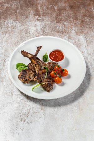 Photo for Three pieces of grilled lamb rack lie on a light ceramic plate. Nearby are cherry tomatoes, lettuce and a gravy boat with adjika. The plate stands on a light fabric background. - Royalty Free Image