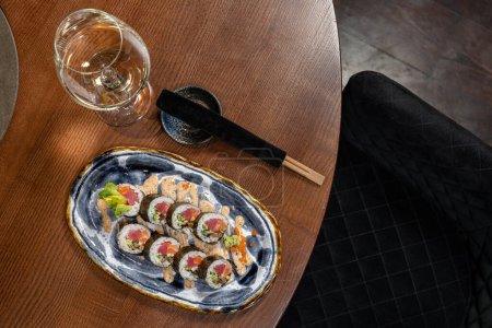 Foto de Rollo de sushi con salmón y atún envuelto en arroz y nori. Se encuentra en un plato de cerámica con un patrón azul en una mesa de madera. Cerca hay una copa de vino blanco y palillos en una caja. - Imagen libre de derechos