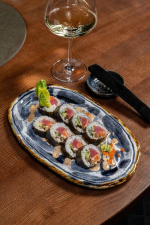 Téléchargez les photos : Rouleau de sushi avec saumon et thon enveloppé dans du riz et nori envahi par la végétation. Il repose sur une plaque en céramique avec un motif bleu sur une table en bois. A proximité se trouve un verre de vin blanc et des baguettes dans une caisse. - en image libre de droit