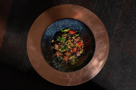Photo for Soba noodles with veal and mushrooms, paprika, broccoli and microgreen sprouts in a dark ceramic plate. The plate is on a copper tabletop. - Royalty Free Image